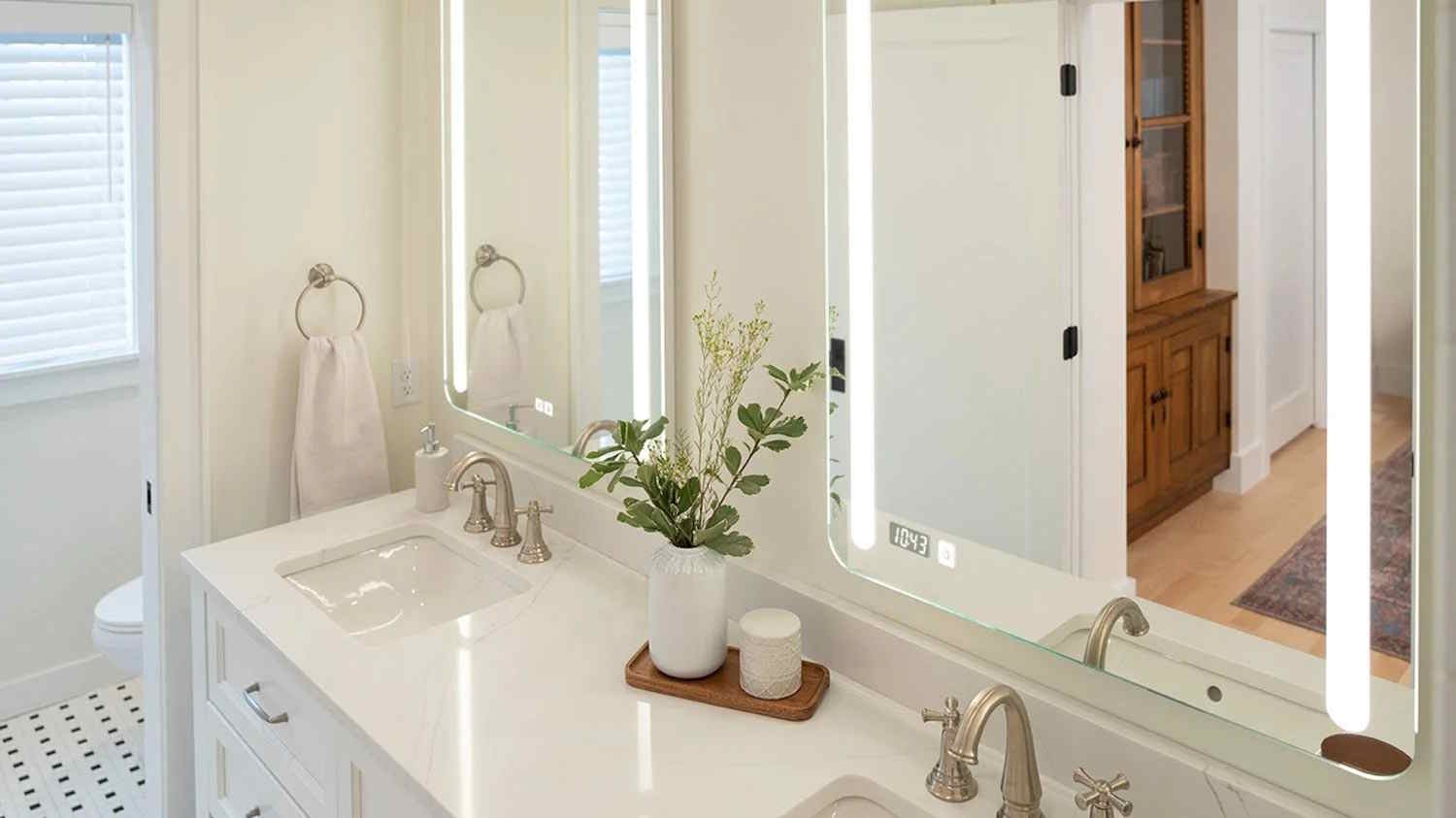A modern double-sink vanity with LED-lit mirror, brushed nickel fixtures, and minimalist decor including a vase with greenery and a candle on a tray.
